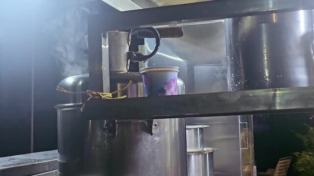 A hazy, close-up shot of a stainless steel tea-making machine, likely in a Kerala restaurant. A container on the machine holds a vibrant, boiling liquid. Steam rises, creating a soft, blurry atmospher