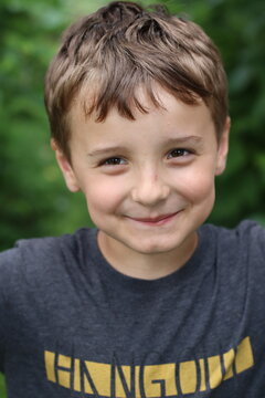 A young boy with short blond hair standing in front of green foliage. He is wearing a grey t-shirt .