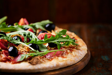 Pizza with salami, tomatoes and mozzarella cheese, black olives and arugula. Rustic wooden background. Soft focus. Copy space. 