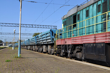 Obraz premium Freight train with a green diesel locomotive and blue cargo wagons at a railway station on a sunny day. Industrial transport concept. 
