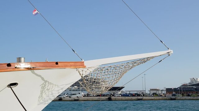 White luxury sailing yacht with traditional rope net bowsprit docked in marina harbor.Beautiful sailing boat bow with rope netting and wooden mast, modern marina with cruise ships in background. 