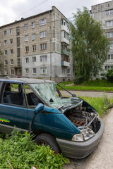 Abandoned blue minivan with smashed windshield and exposed engine parked in overgrown grass near...