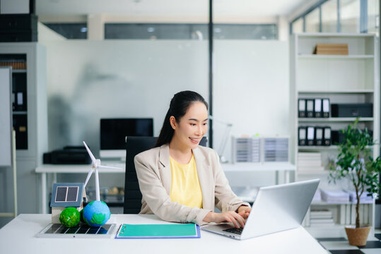 Asian businesswoman in office planning green energy with wind turbine, solar panel, globe. Concept of sustainability, climate change, and reducing CO2 emissions. - Powered by Adobe