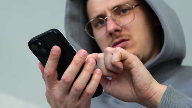 Young guy in glasses and hoodie typing on his touchscreen smartphone.
Using mobile phone to send important message and communicate with people online.
Typing, chat, browser