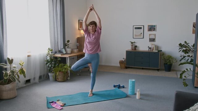 Full shot of slim senior 60-year-old Caucasian woman in pink t-shirt and blue leggings standing on fitness mat at home, practicing yoga in vrikshasana pose, during home fitness routine