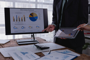 Business team analyzing sales data on computer and tablet, discussing revenue and customer overview, pointing at charts and graphs during a collaborative meeting