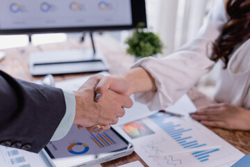 Business partners shaking hands over financial charts, making a deal after successful negotiations in modern office, concept of collaboration and partnership