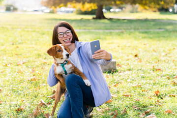 Dog and woman taking selfies with smartphone in autumn park. Girl and pet happy together outdoor....
