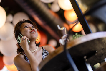 Smiling woman enjoying a phone conversation in a lively cafe with bright lights and a delicious dessert on the table