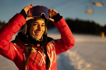 Smiling woman in pink jacket adjusting ski goggles at sunset in snowy landscape