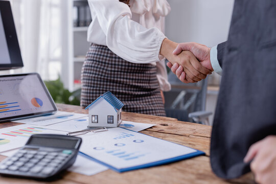 Real estate agent and client shaking hands after finalizing a contract in a professional office setting, surrounded by a house model, calculator, documents, and a tablet on the desk