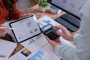 Business colleagues are collaborating on sales performance analysis, using a digital tablet and calculator to review charts and graphs, working together to assess financial data