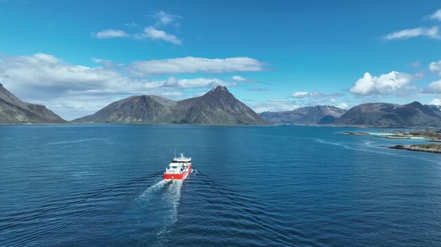 Well boat for salmon farming sails past Gimsoy Island in the Lofoten archipelago, Norway, traveling upstream, slow motion aerial, following behind boat.