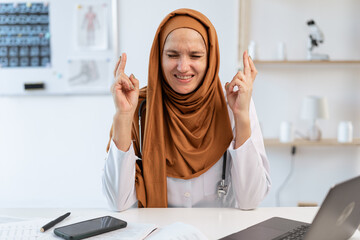 Hopeful Muslim woman doctor cross fingers and smiling, making wish and praying working at desk in...