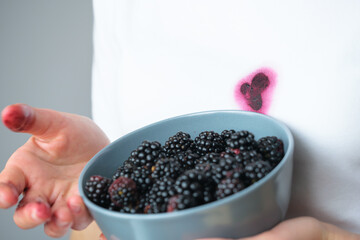 A child stands indoors, holding a bowl filled with ripe blackberries. Stains on their shirt hint at...