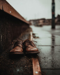 Brown shoes rest close together by wet curb on rainy city sidewalk; shoes look weathered as brown shoes suggest solitude and passage of time in moody urban scene