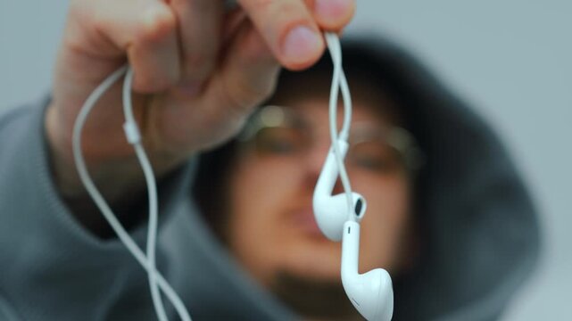 Modern European guy in gray coffee holds wired headphones in his hands in the foreground.
Faded wired headphones for listening to music and talking on the phone.