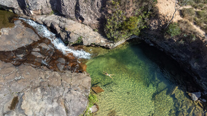 Drone shot of a person relaxing in a crystal-clear natural pool surrounded by rocky cliffs and a small waterfall. Peaceful travel and nature concept
