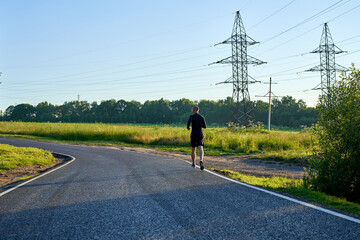 A runner athlete runs along a rural road among fields and forest in the countryside. View of the...