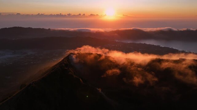 A golden sunrise cast light over Mount Batur in Bali, Indonesia, with hikers gathered at the summit to witness the mist veiling volcanic peak and glowing clouds.