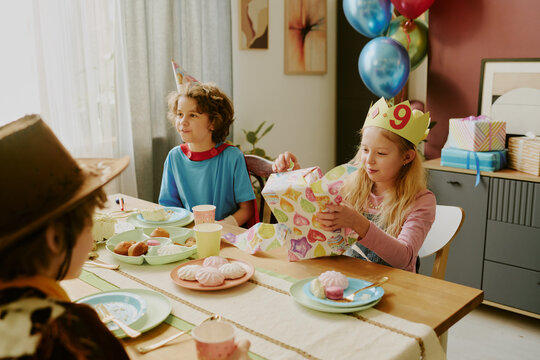 Caucasian girl child wearing birthday crown opening present while sitting at table with two other children during birthday party, colorful balloons and wrapped gifts in background - Powered by Adobe