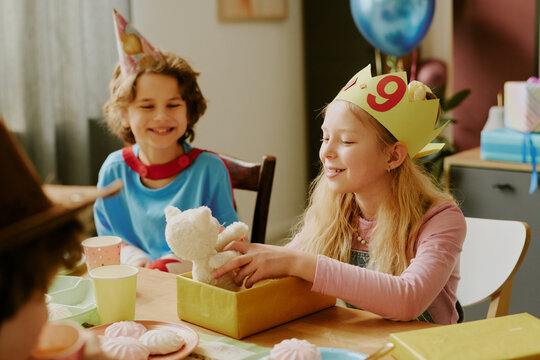 Caucasian girl wearing birthday crown with number nine smiling while opening gift box containing stuffed animal, sitting at table with other children during birthday celebration