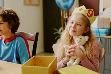 Caucasian girl child wearing paper crown with number nine smiling and hugging stuffed animal, while...