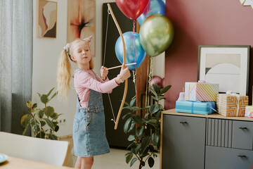 Caucasian girl child wearing party hat holding toy bow aiming at colorful balloons, standing near...
