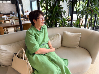 Middle-aged woman in green dress sitting on sofa with handbag. Lifestyle, leisure, and modern...