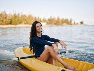 A young woman sits in a yellow kayak on a sandy shore, smiling softly as gentle waves lap the lake....