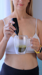 Woman preparing healthy matcha latte whisking green tea in clear glass mug
