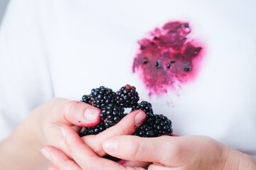 A hand grips ripe blackberries, their juice staining fingertips and a white shirt nearby. The...