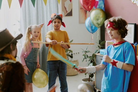 Group of children including Caucasian girl and boys of diverse ethnicities playing with toy swords and balloons during indoor party, kids wearing costumes and smiling while interacting - Powered by Adobe