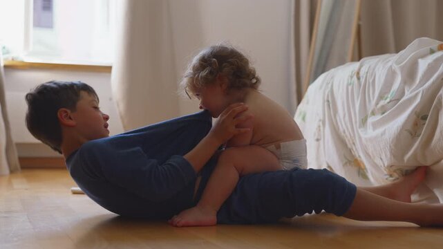 Young boy leaning forward as toddler playfully pulls on his shirt with teeth during fun sibling interaction on floor at home