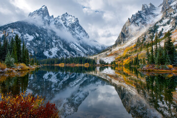 Tranquil lake reflecting snow-covered rugged mountain peaks and colorful autumn foliage under a cloudy sky in a serene wilderness landscape