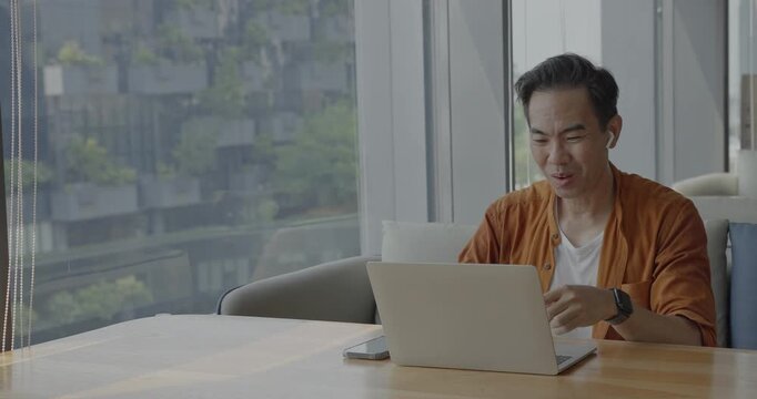 Senior Asian businessman wearing headset celebrates success during online meeting in modern co-working space. Remote work and business communication concept	
