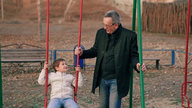 Happy family. Little son smiling sits on swing, dad swings child on children's swing in autumn park. Little son plays on swing on playground under supervision of parent dad on playground autumn park - Powered by Adobe