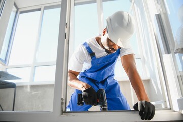 Workman in overalls installing or adjusting plastic windows in the living room at home