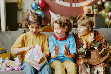 Three diverse children sitting on sofa celebrating birthday, wearing party hats, one child opening gift box while two friends watching and clapping, colorful balloons in background