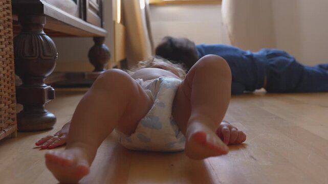 Toddler lying on floor in diaper reaching hand toward older brother during playful sibling bonding activity and fun interaction at home