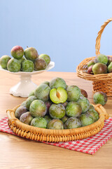 Fresh Green and Red Plums in Wicker Baskets on Wooden Table with Cut Fruit Showing Yellow Flesh