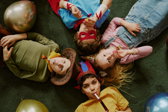Group of four children lying on floor blowing party horns, two boys and two girls including Caucasian and multiethnic kids wearing costumes and accessories during celebration - Powered by Adobe
