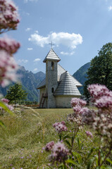 A stone chapel stands peacefully in a green meadow, surrounded by towering mountains under a blue sky, framed by soft pink flowers in the foreground in the village of Theth in Albania