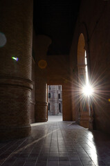 Sunlight bursts through a grand brick archway, creating a dramatic starburst effect and casting long shadows along a tiled corridor, highlighting the building's texture and depth.