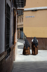 Two nuns in traditional habits walk down a narrow, sunlit street in a historic Spanish city. The image captures a timeless moment, contrasting the traditional religious attire 