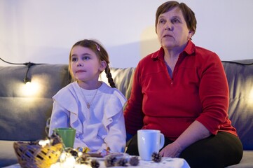 A young girl and her grandmother relax on a couch, illuminated by warm string lights, sharing a...