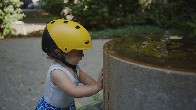 Toddler touching stone fountain outdoors wearing yellow helmet, observing bubbling water with curiosity, playful summer moment of exploration and early discovery