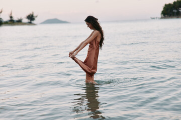 Woman standing in shallow sea water wearing a flowing dress, enjoying peaceful sunset with calm...