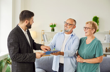 Real estate agent gives contract to smiling senior couple at home, explaining agreement and...