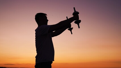 Adult man exercising outdoors. Motivation to exercise active lifestyle. Man doing strength training, lifting dumbbells in front of him outdoors. Man exercising with dumbbells in park sunset. Fitness